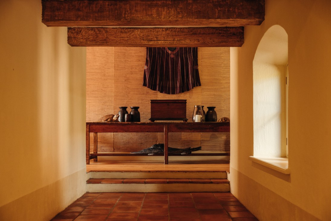 Interior hallway with exposed wooden ceiling beams, woven wall panel, traditional garment display and terracotta floor