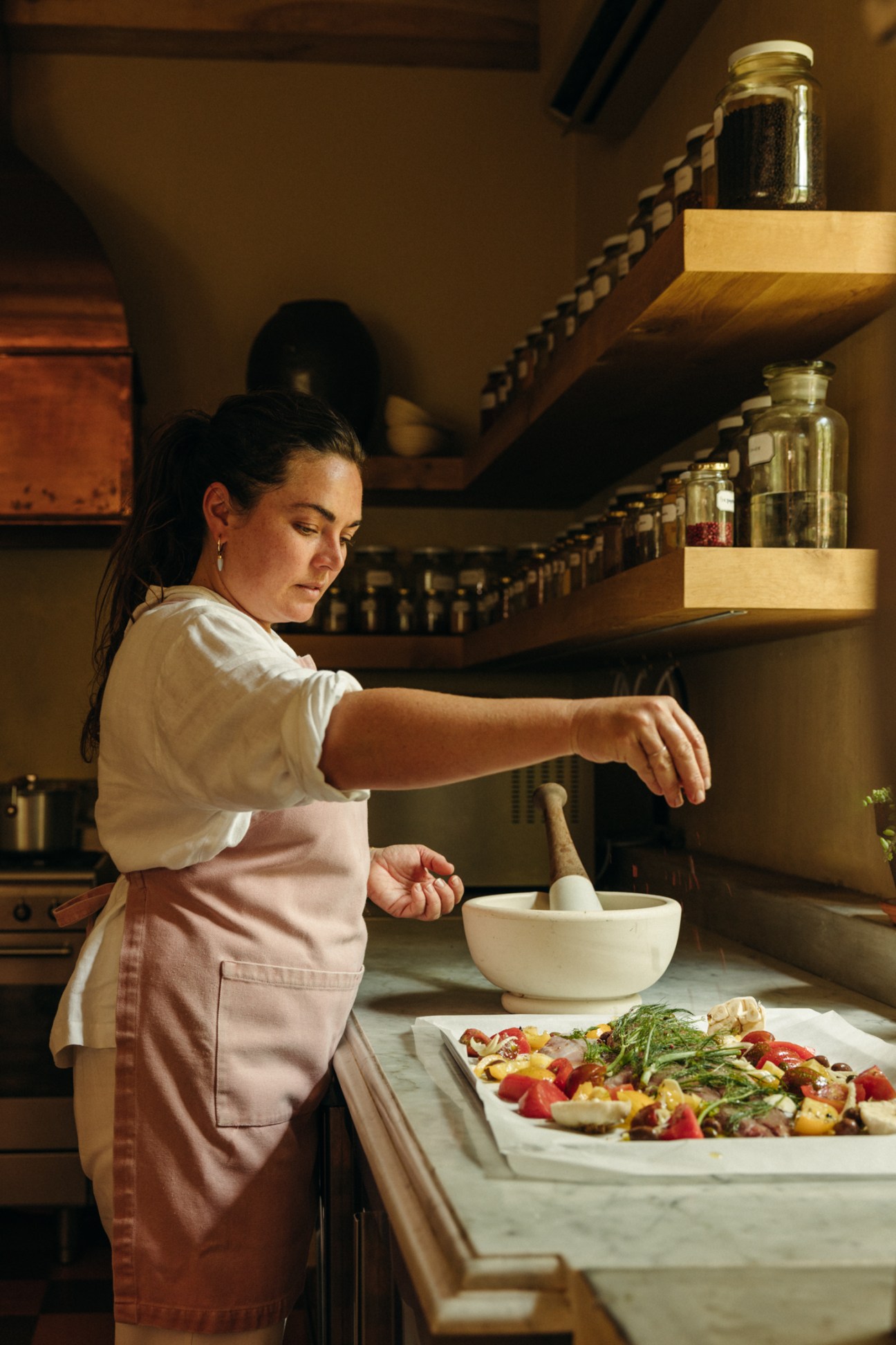 Woman in white shirt and pink apron using mortar and pestle in rustic kitchen with jars on wooden shelves