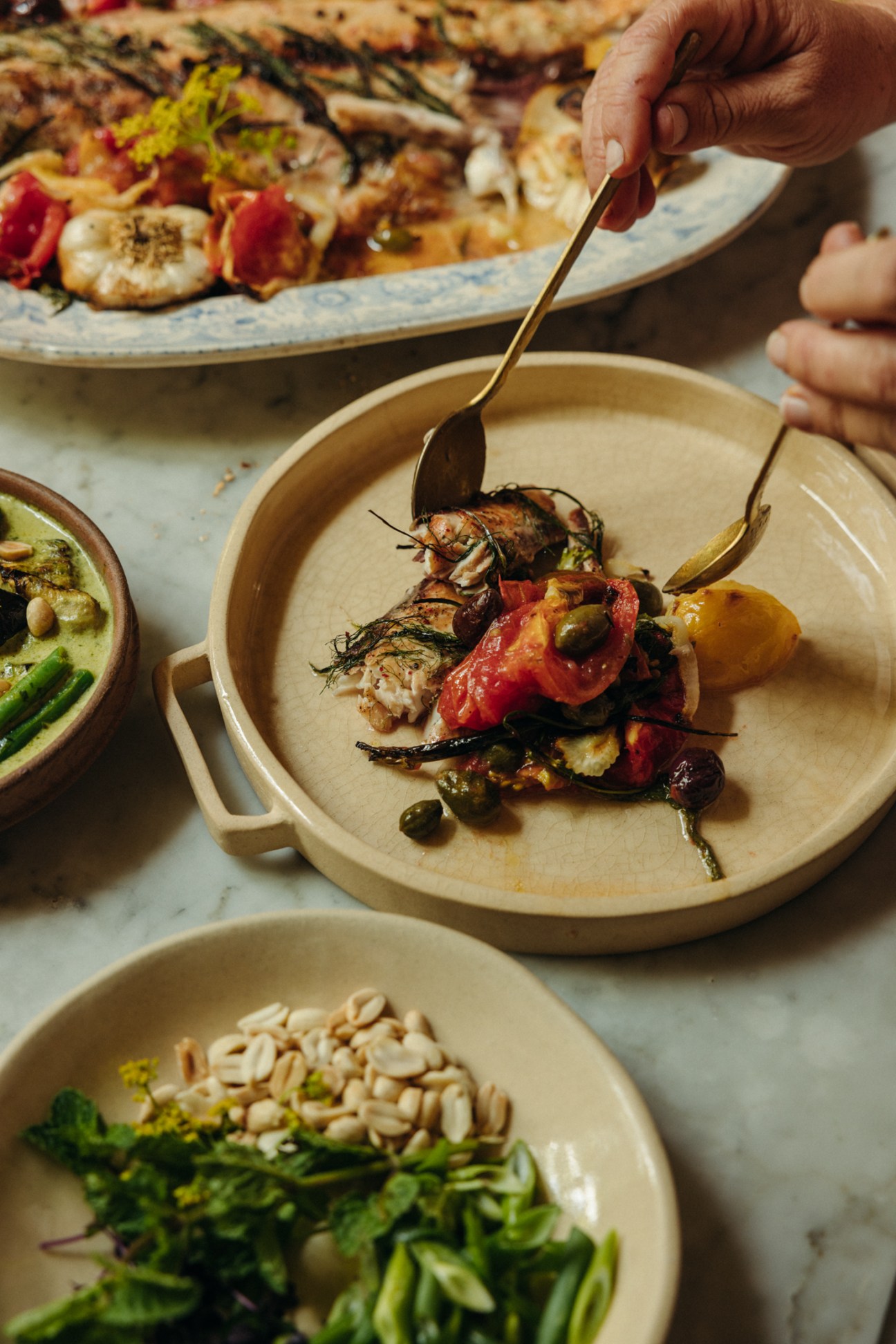 Hand holding spoon over ceramic bowl with grilled vegetables and garnishes, with plates of food in background