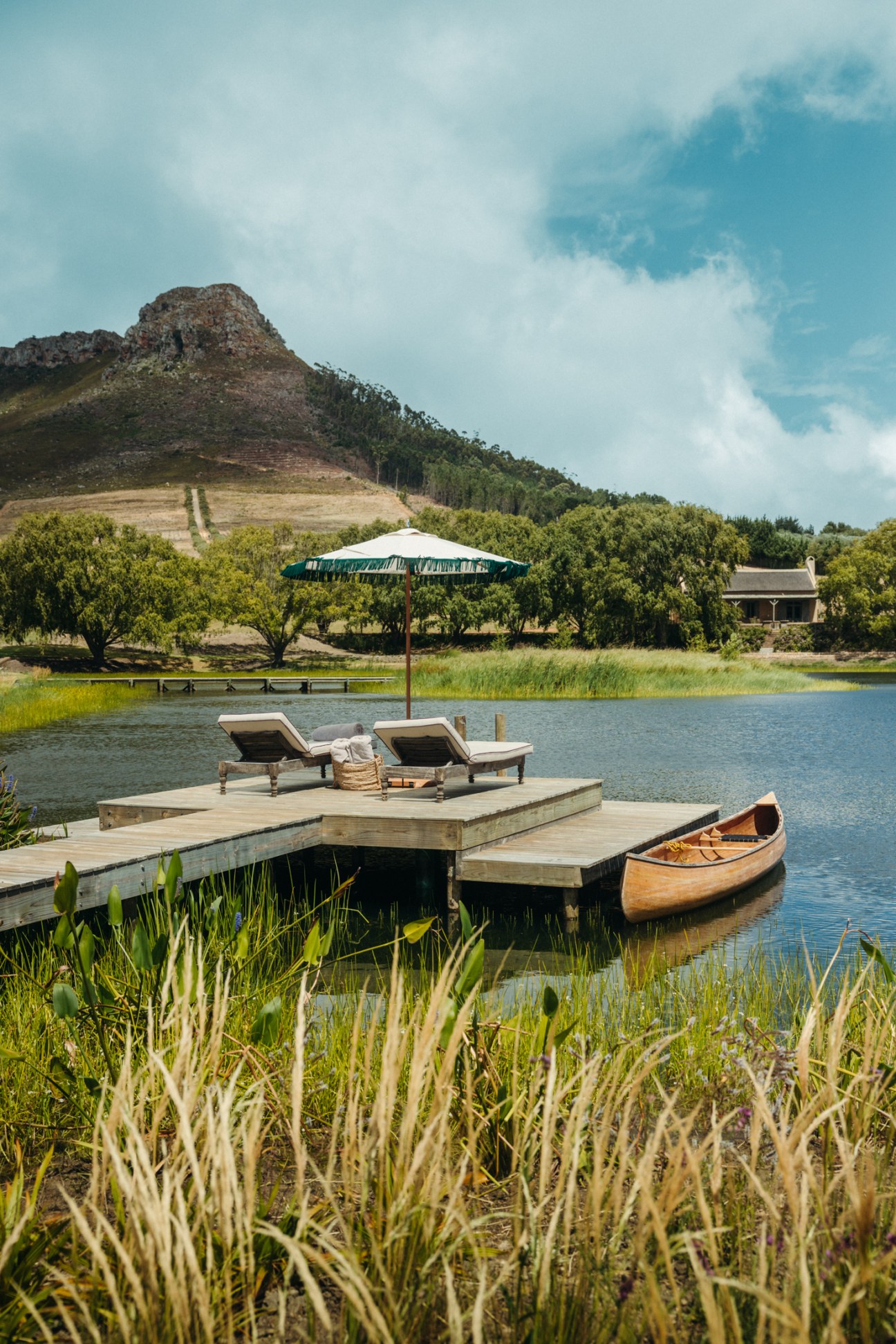 Wooden dock with two lounge chairs under umbrella, wooden canoe moored alongside, lake and mountains in background