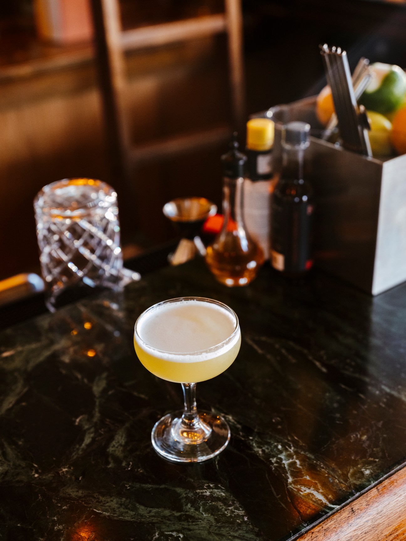 A cocktail in a coupe glass sits on a dark marble bar with bartending tools and glassware in the background