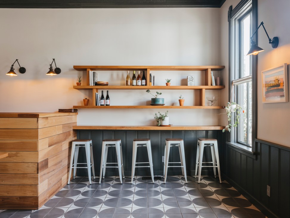 Modern kitchen bar with white metal stools, wooden shelving displaying bottles and books, and geometric patterned floor