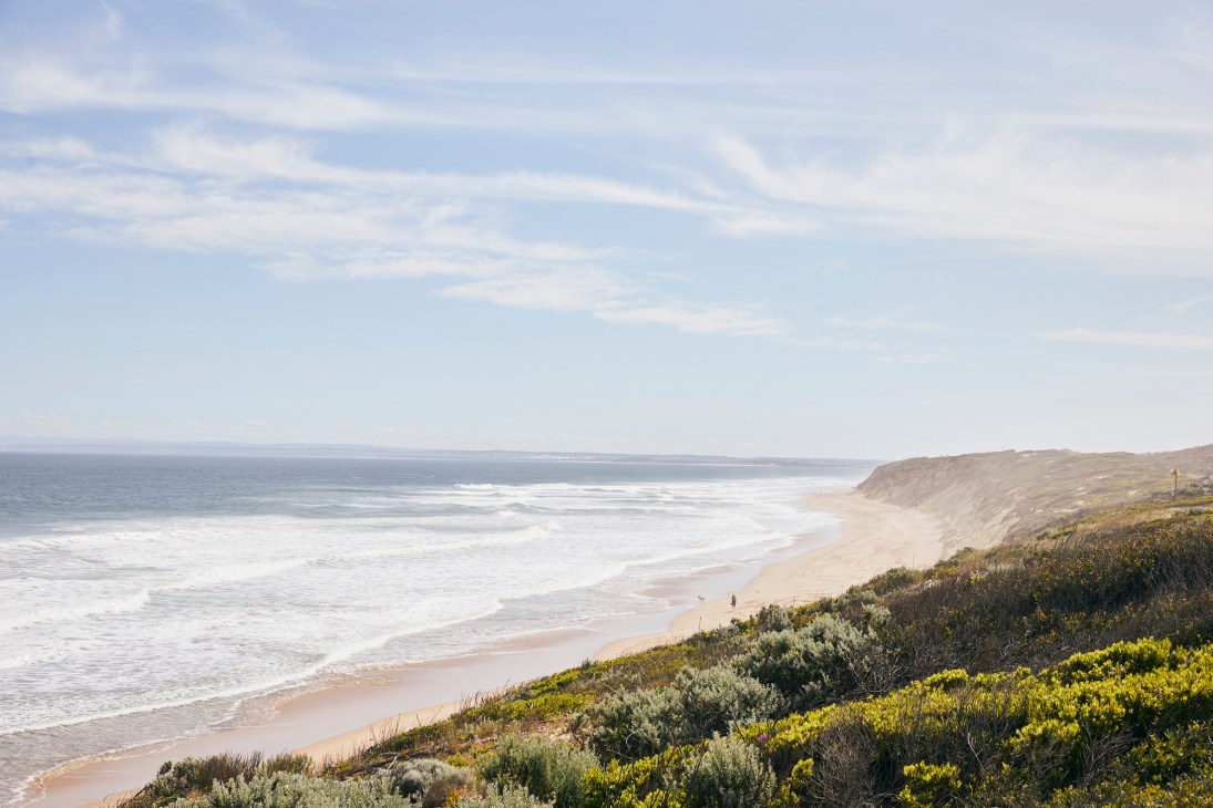Coastal beach landscape with sandy shore, ocean waves, and vegetated cliffs under blue sky.