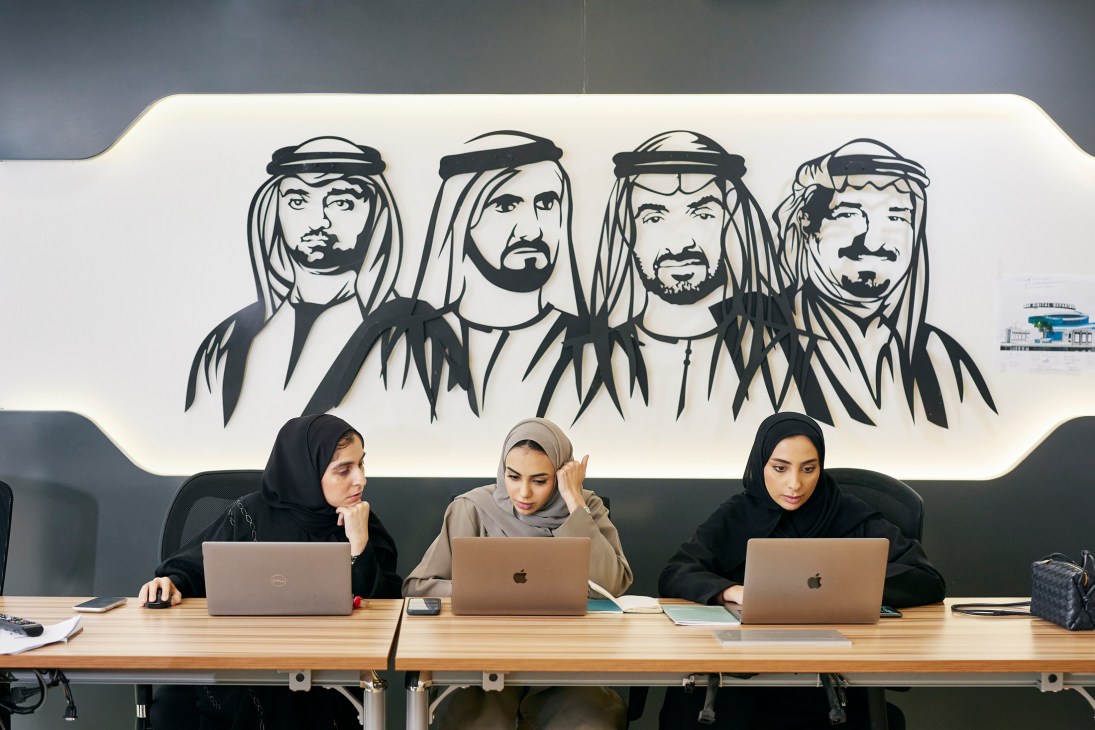Three women in headscarves work at laptops at a wooden desk beneath a mural of four men in traditional Arab dress.