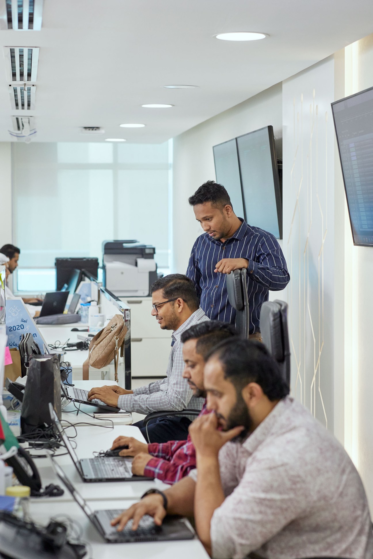 Multiple people working at computer terminals in a modern office with white walls and recessed lighting.