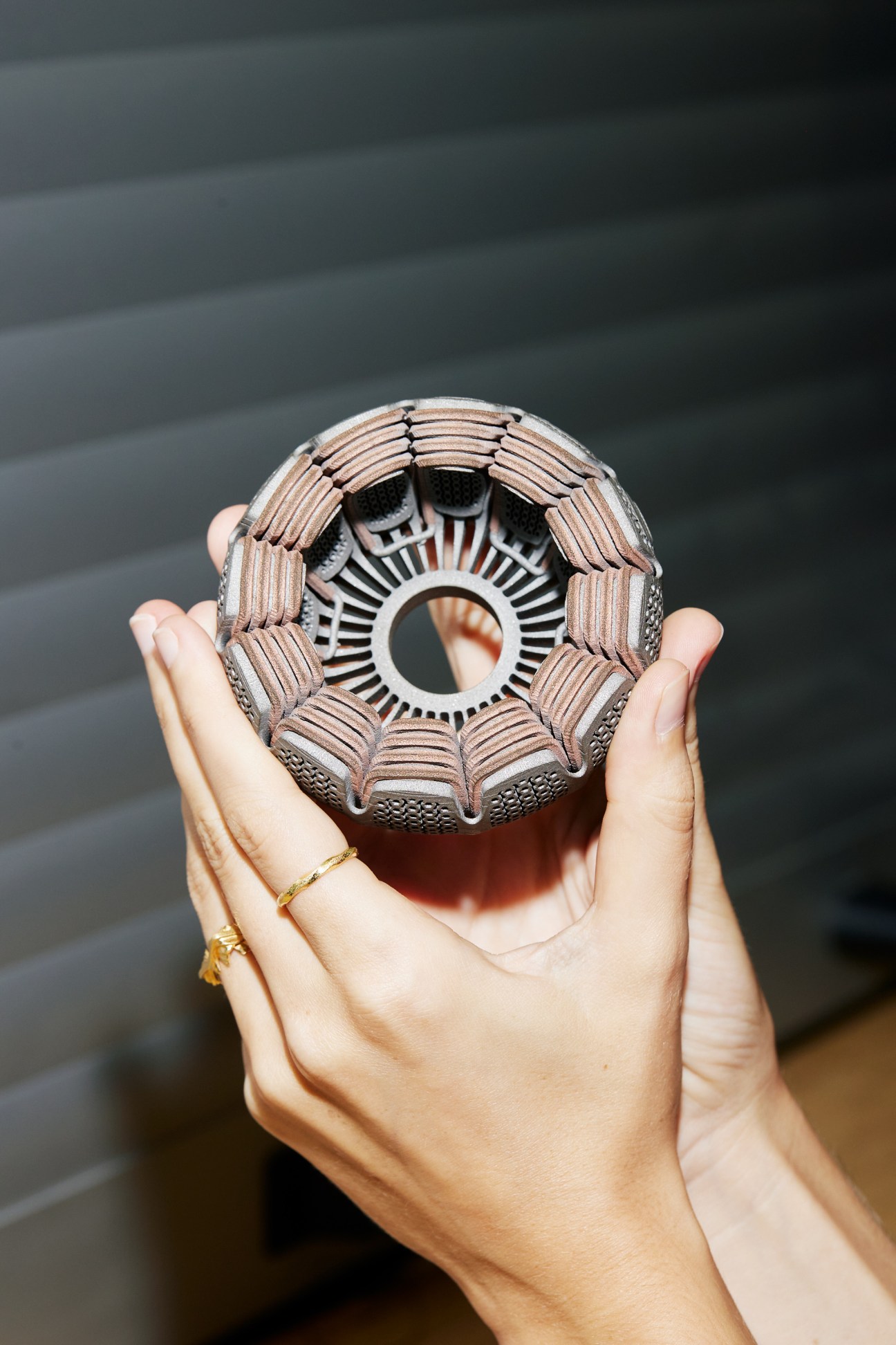Hand holding a 3D-printed electric motor coil with intricate copper-colored ribbed patterns and central opening.