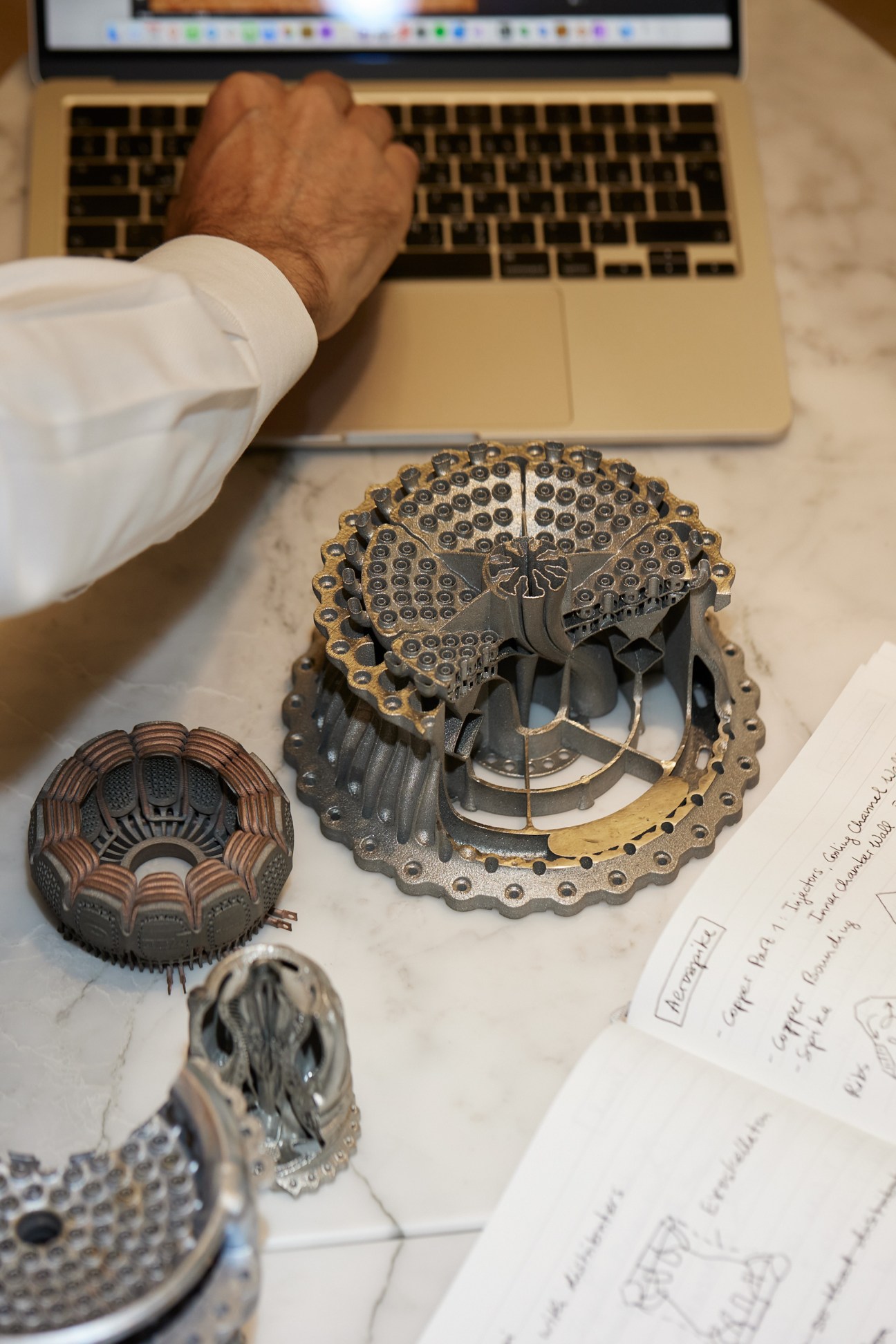 3D-printed metal engine parts displayed on a marble table next to a laptop and design sketches.