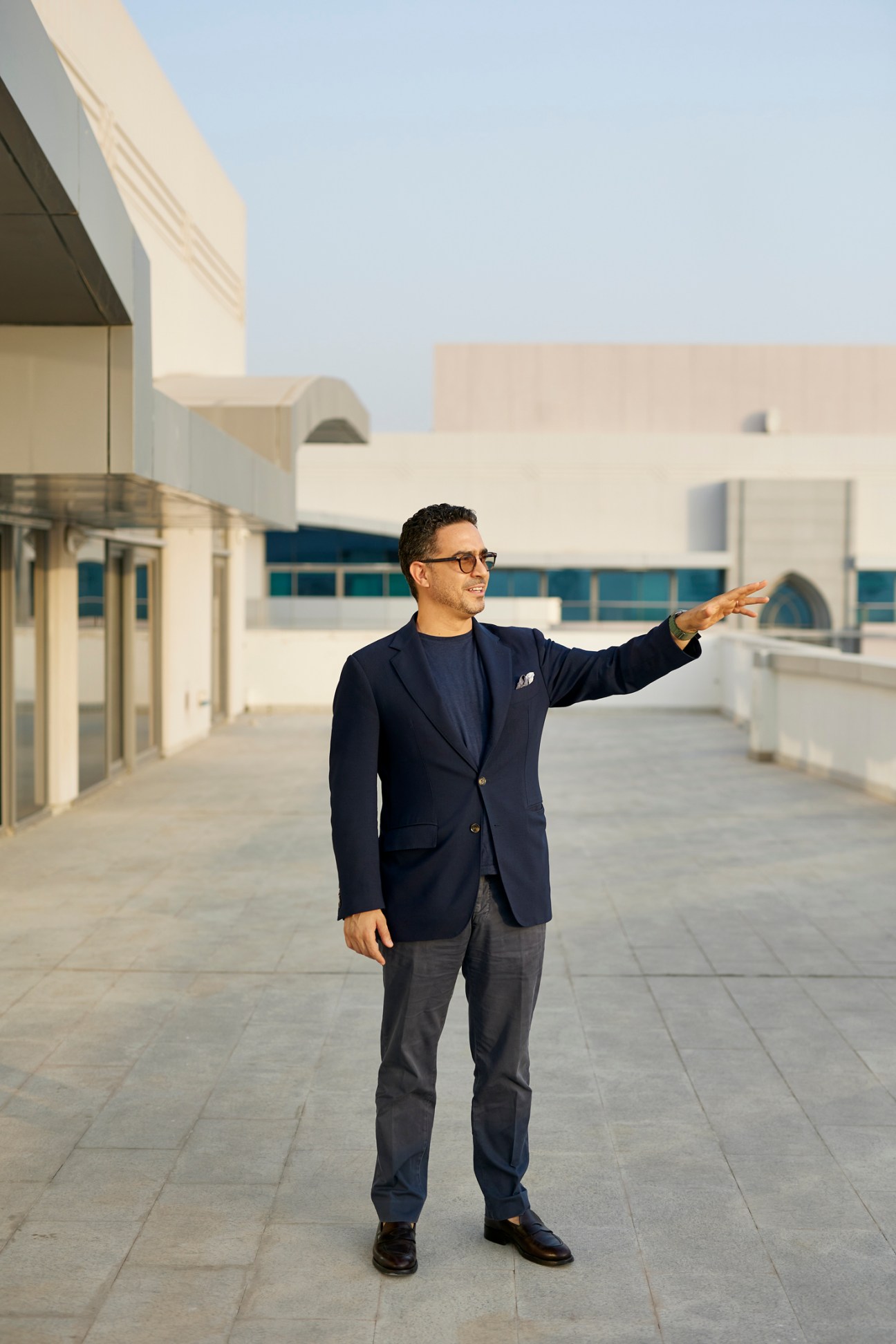Man in navy blazer gesturing toward modern white buildings on a rooftop in Abu Dhabi.