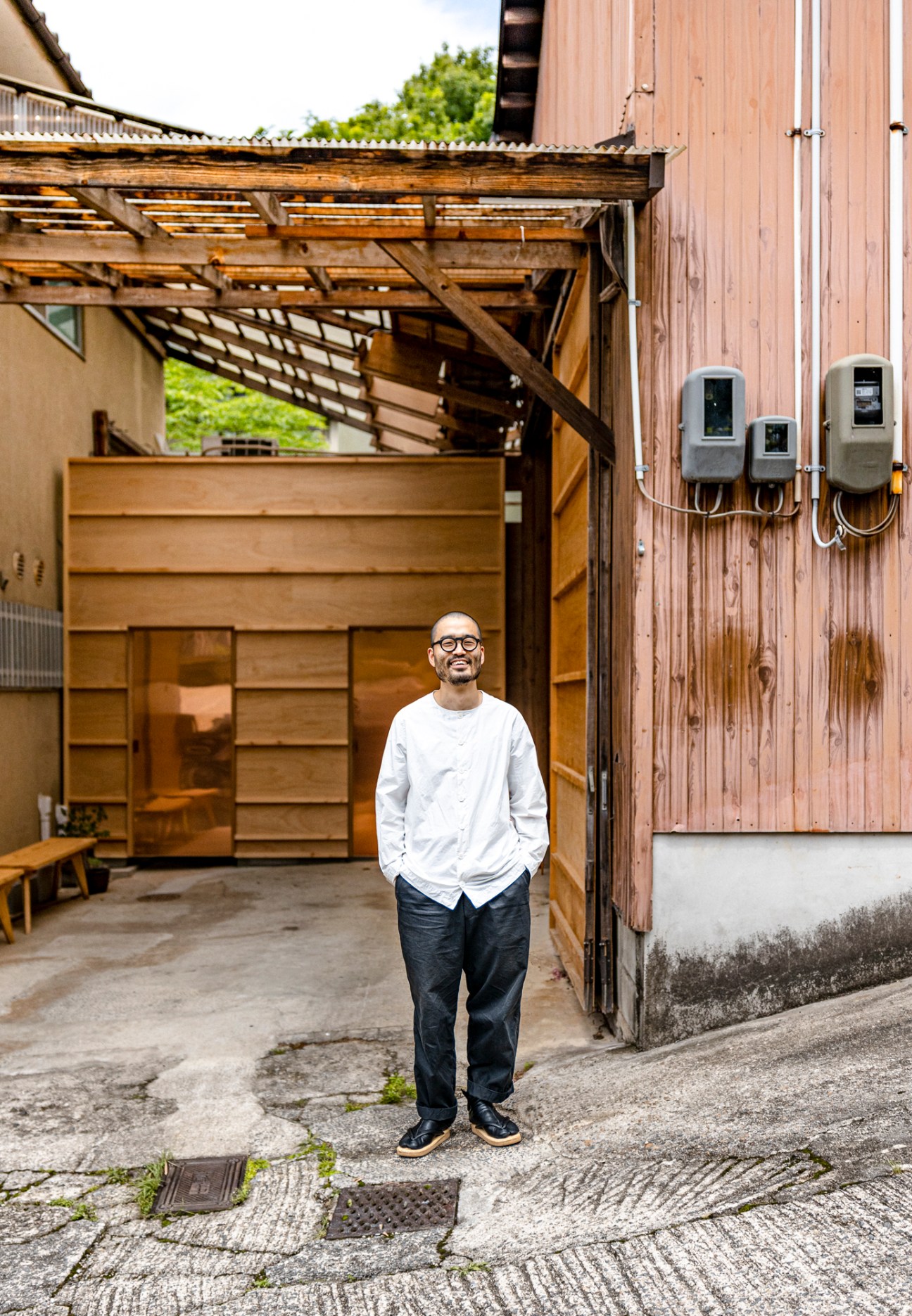 Shinji Sekizuka stands in the courtyard of his timber warehouse atelier in Kyoto, wearing glasses, white long-sleeve shirt and dark trousers.