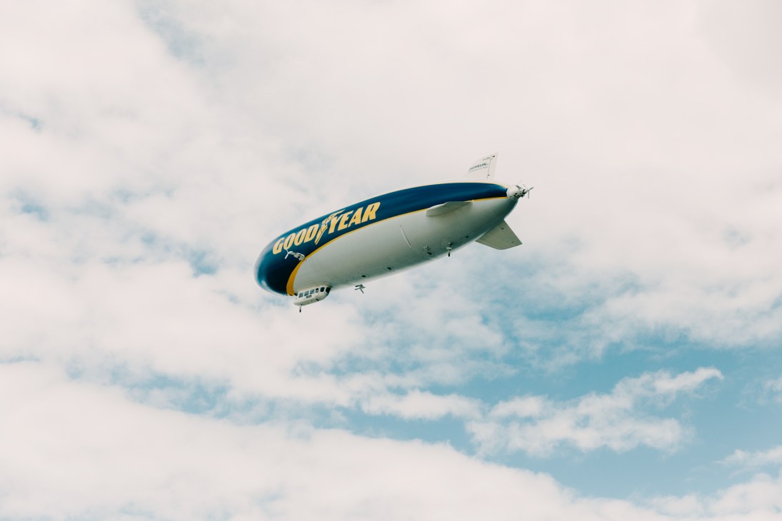Goodyear blimp flying through cloudy sky with blue and yellow branding visible