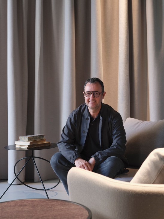 A man wearing glasses and a dark jacket sits on a beige sofa in a minimalist interior space with floor-to-ceiling curtains, with a small black side table holding stacked books beside him.