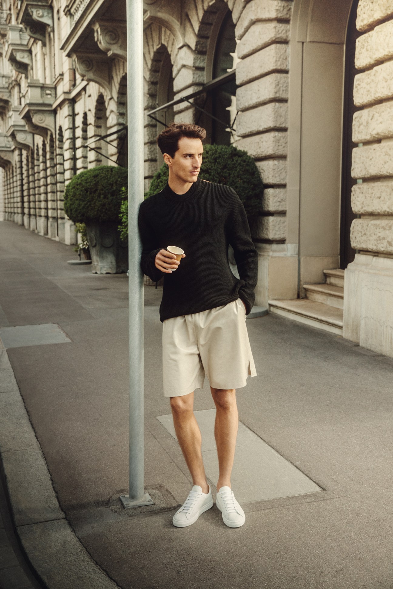 Man in black Bottega Veneta jumper and beige shorts with white Pelikamo trainers holding coffee on urban sidewalk