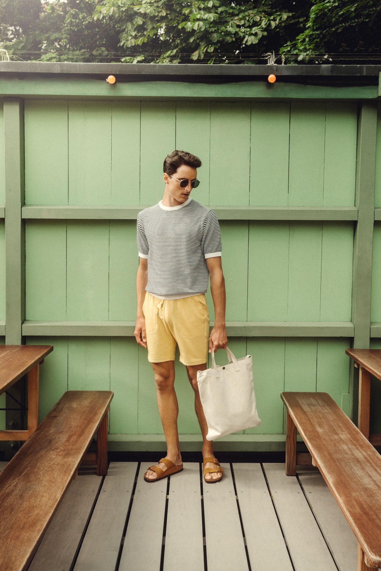 Man in striped jumper and yellow shorts holding white bag, wearing sunglasses and sandals on wooden deck