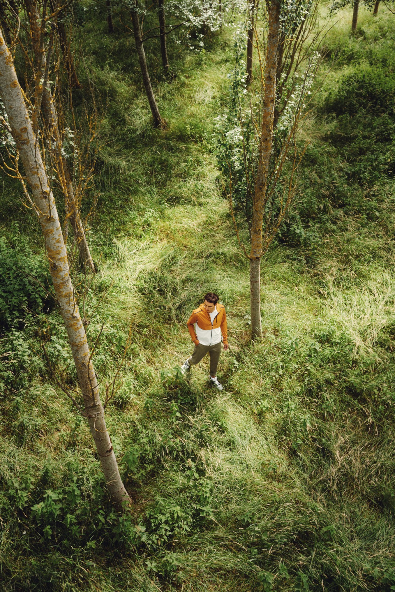 Person in orange and white jacket walking through lush green forest trail with tall grass and trees