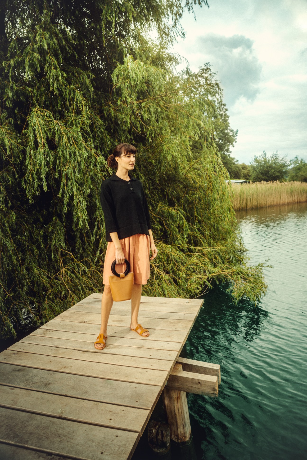 Woman in black pullover and coral shorts with yellow bag standing on wooden dock by water