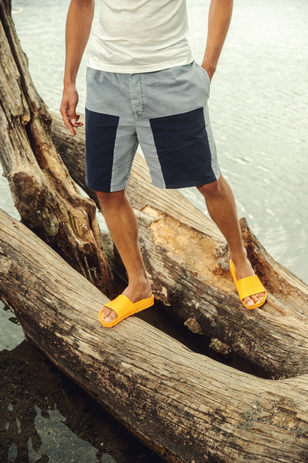 Man in white t-shirt and colorblock shorts with yellow Birkenstock sandals standing on driftwood by water