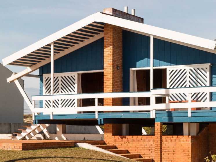 Modernist house with blue wooden siding, white trim, brick chimney, and geometric railings under clear sky