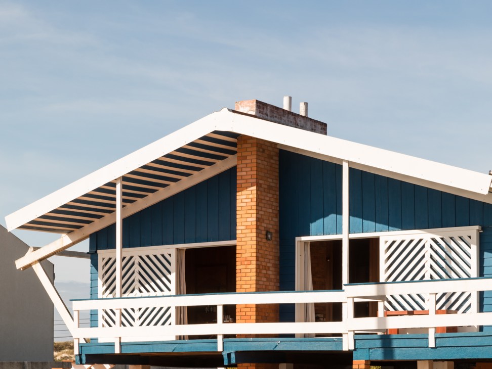 Modernist house with blue wooden siding, white trim, brick chimney, and geometric railings under clear sky