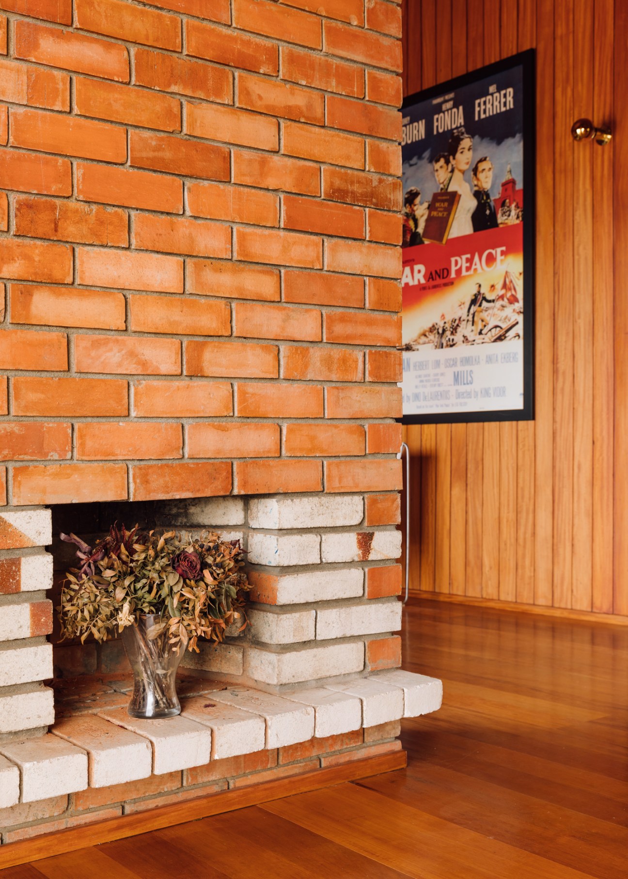 Brick fireplace column with vase of dried flowers in living room with wood paneling and movie poster