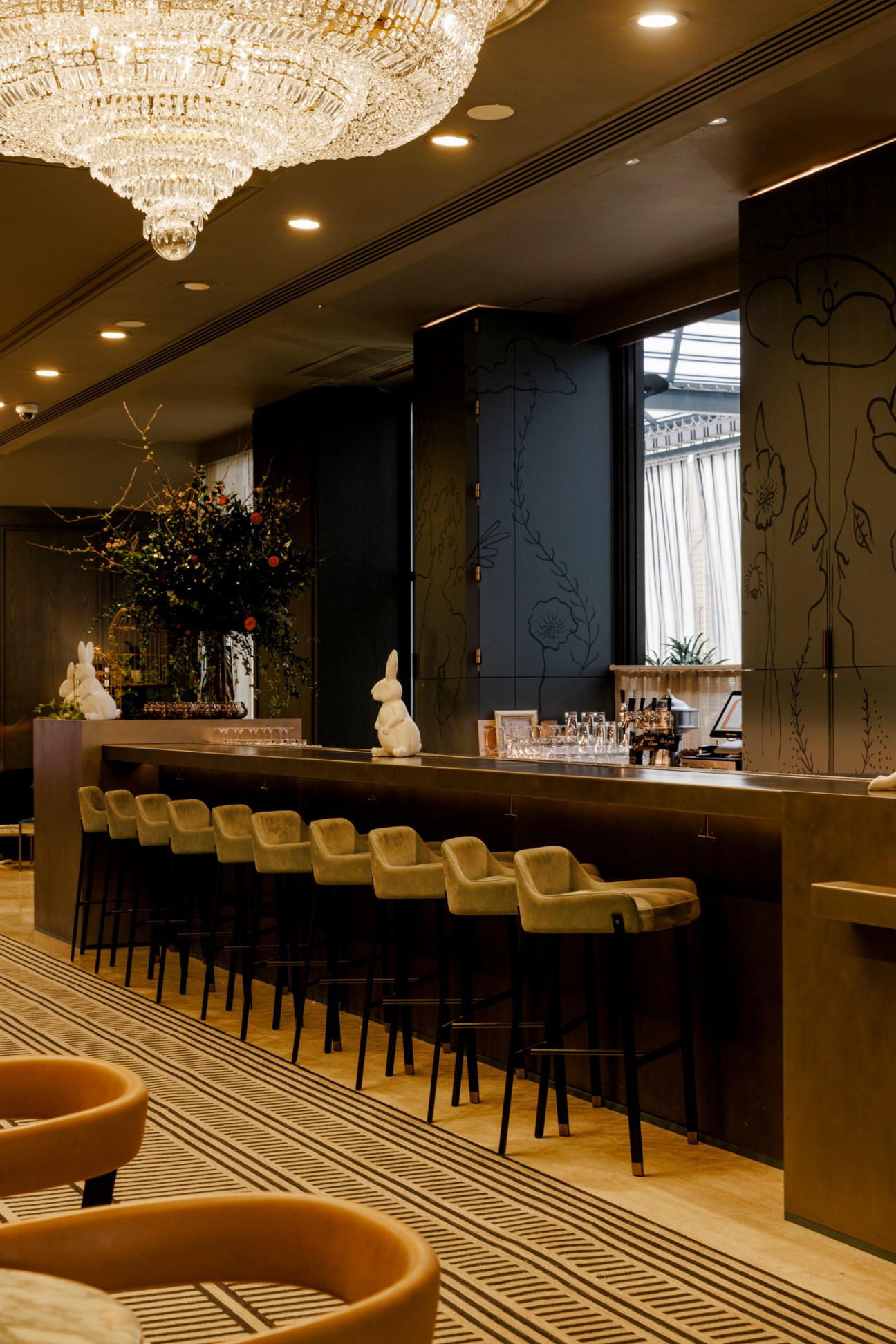 Elegant hotel bar with crystal chandelier, dark wood counter, mustard yellow bar stools, and decorative wall murals