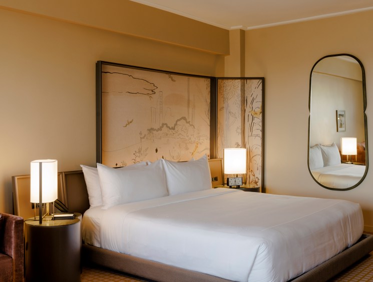 Hotel room with white bedding, artistic headboard panel, warm lighting, and oval mirror on beige walls