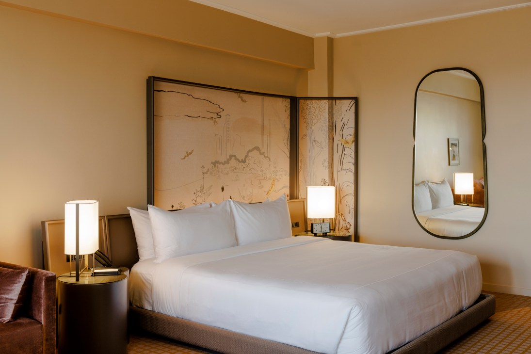 Hotel room with white bedding, artistic headboard panel, warm lighting, and oval mirror on beige walls