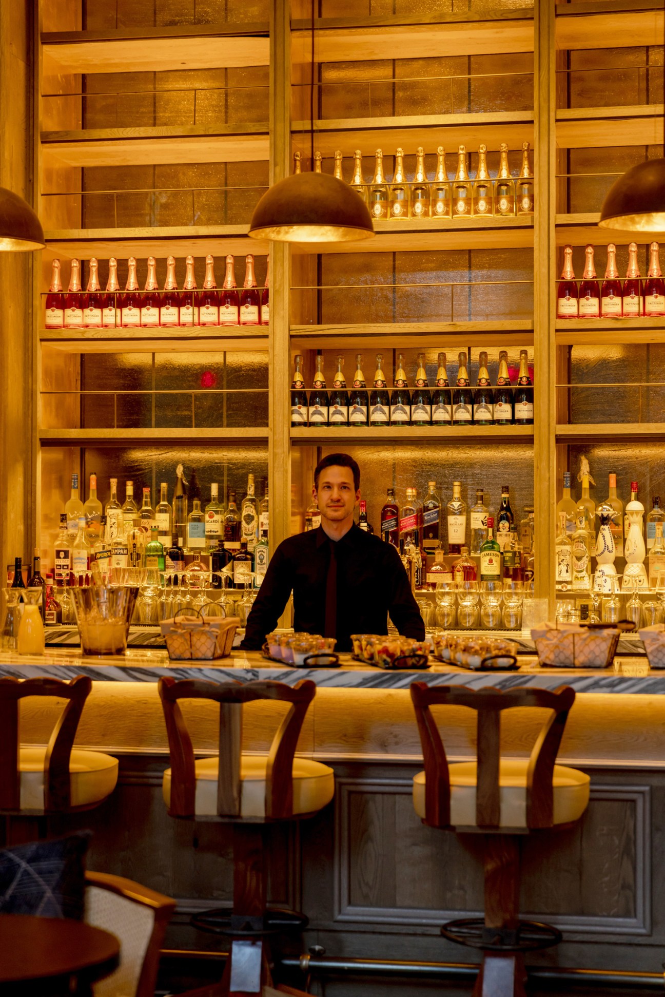 Bartender in black shirt standing behind illuminated bar with extensive liquor collection and pendant lighting