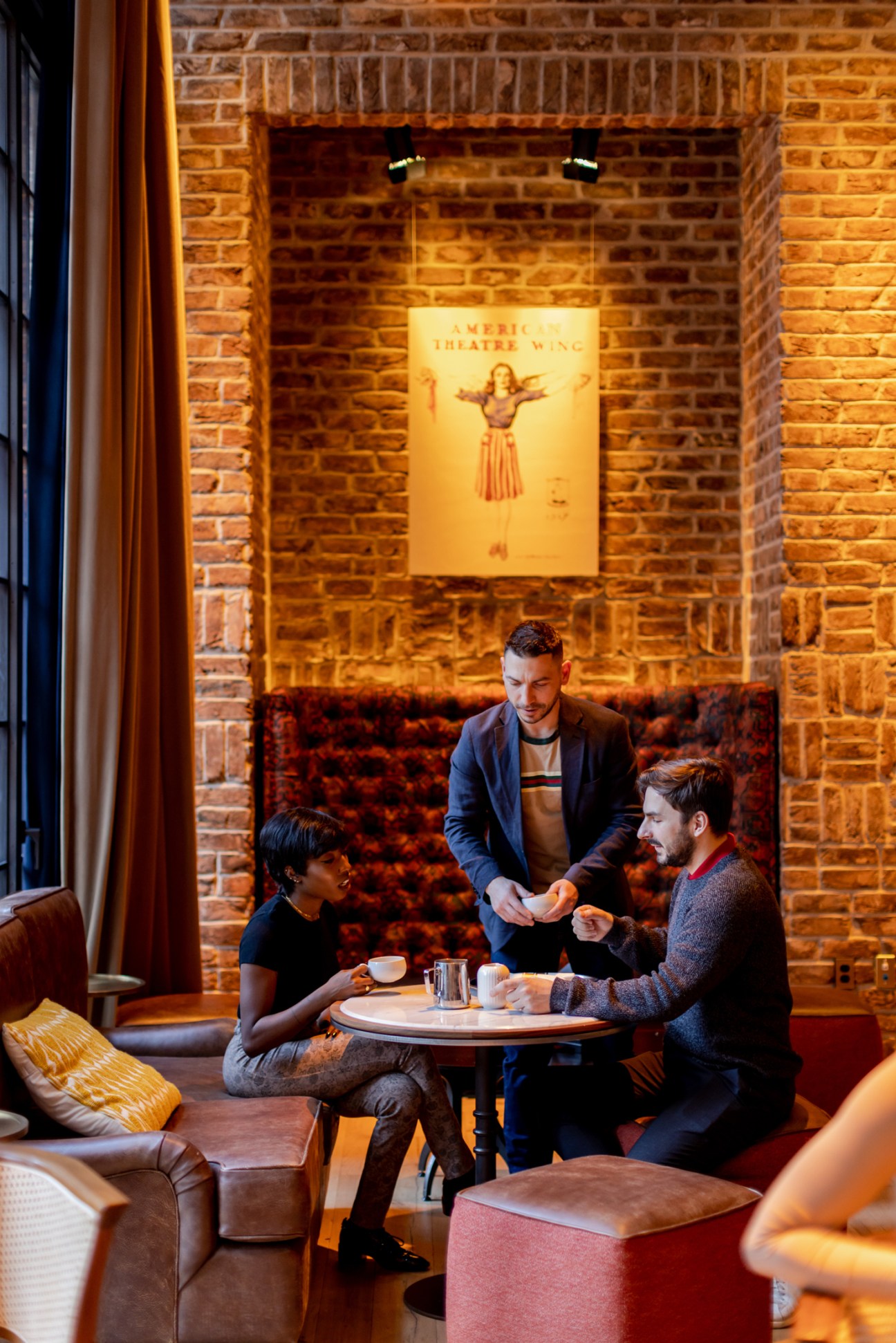Three people in a hotel lounge with exposed brick walls, one server standing while two guests sit at a small table