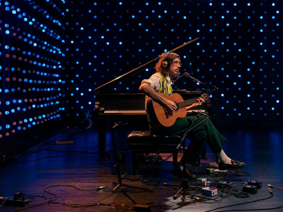 Tim Bernardes performing acoustic guitar in radio studio with microphone and piano visible