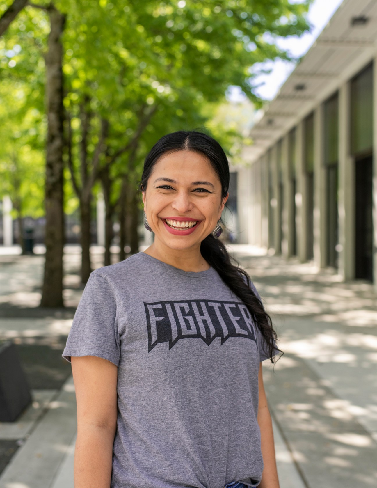 Albina Cabrera in grey 'Fighter' t-shirt standing outdoors under tree-lined covered walkway