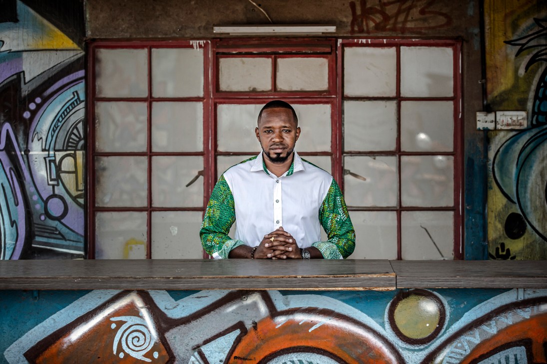 Boniface Mwangi, Kenyan political-party founder, stands at a counter wearing a white shirt and green patterned sleeves against a red-framed window.