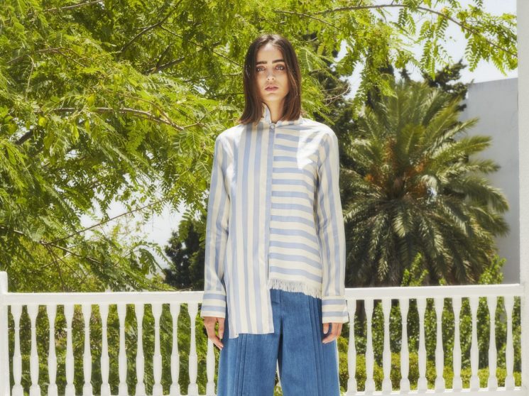 Woman in blue and white striped top with wide-leg denim pants standing on sunny terrace with green foliage