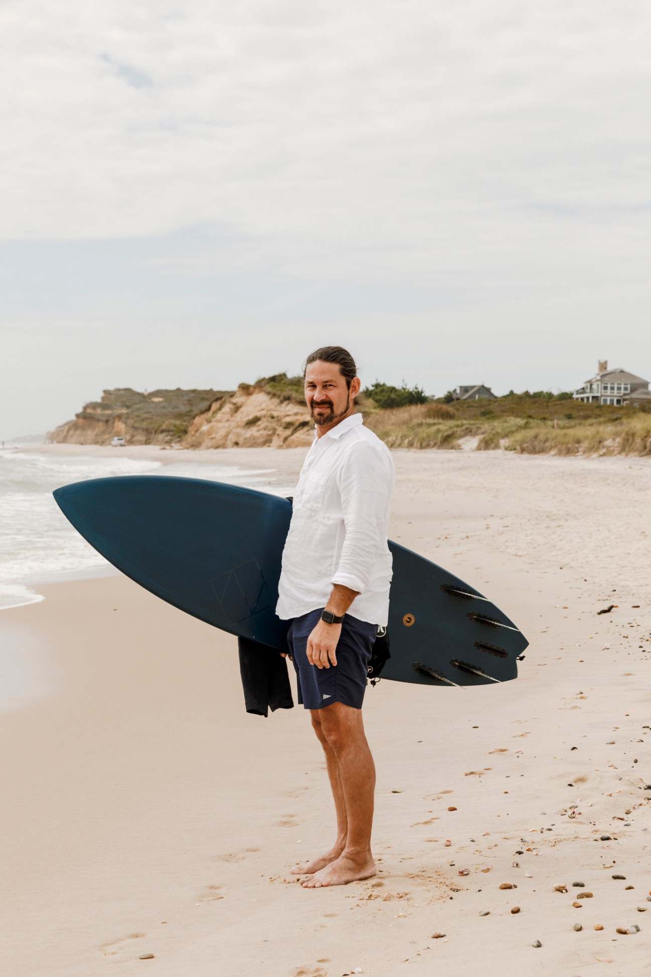 Tommy Zung holding a dark surfboard on a beach with coastal cliffs and houses in the background