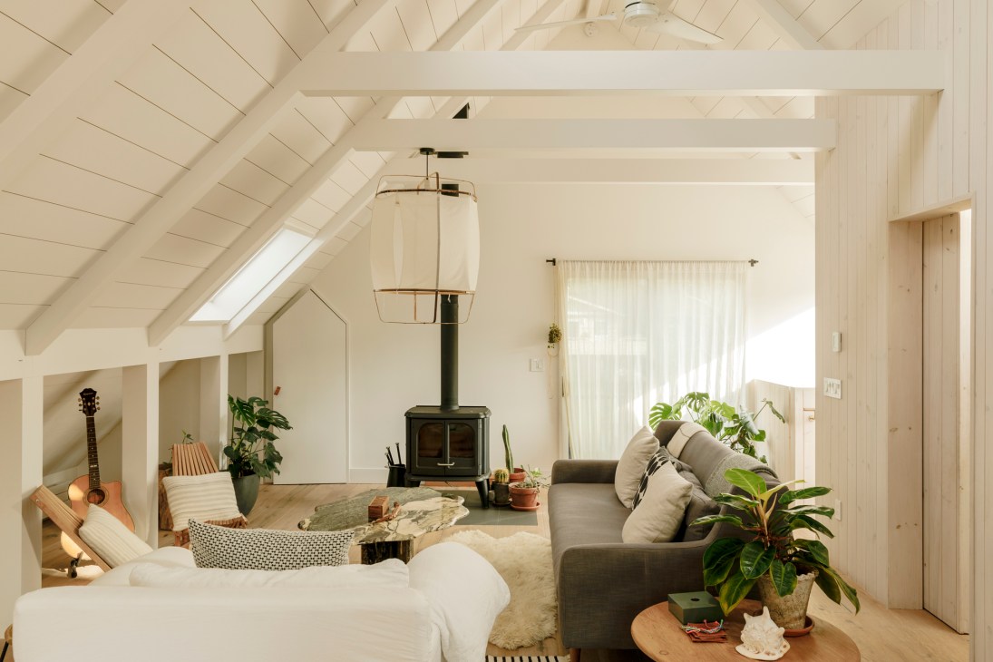 Modern attic living space with exposed white beams, wood-burning stove, gray sofa, and natural light from skylights