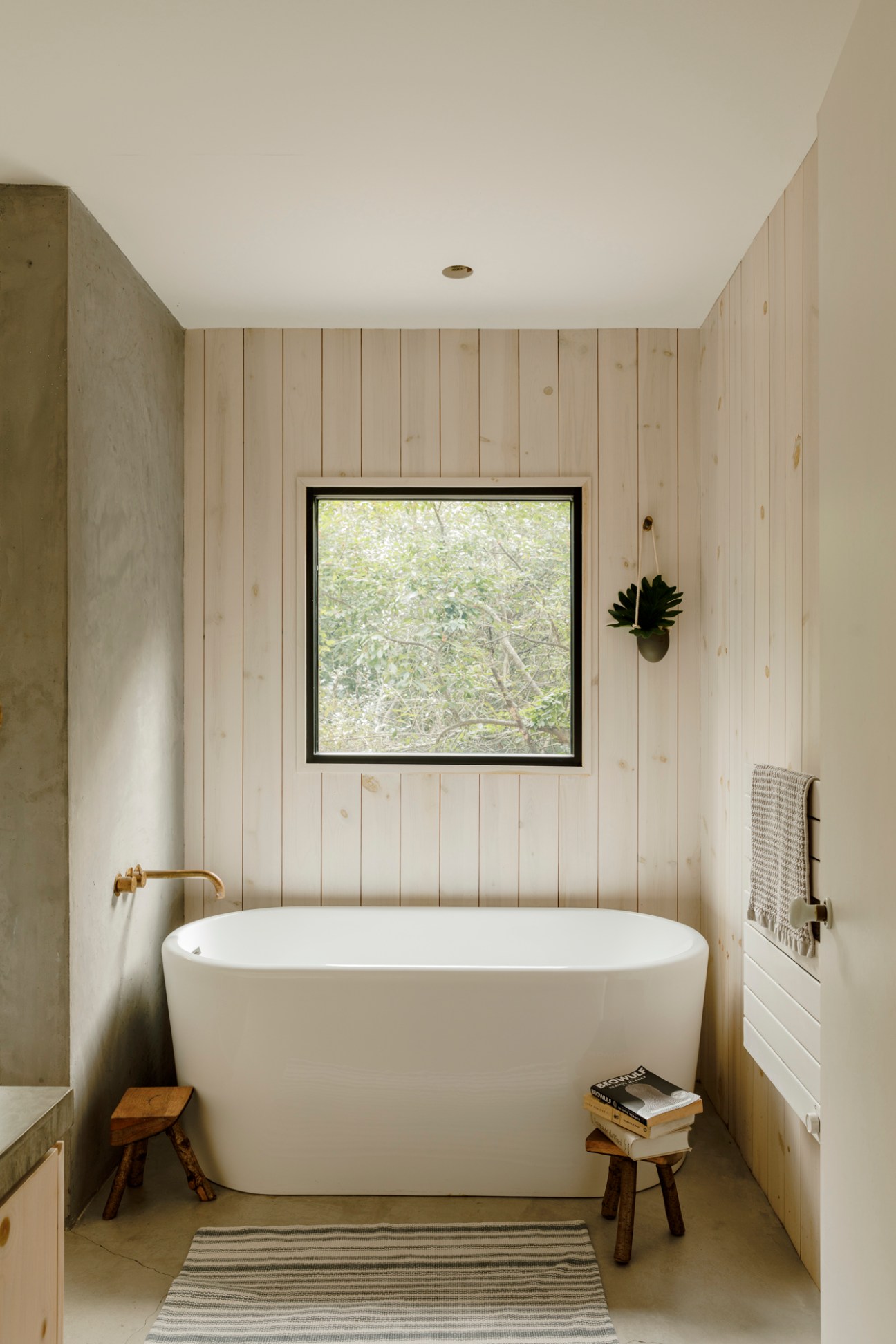 Modern bathroom with white freestanding bathtub, natural wood paneling, and large window overlooking greenery