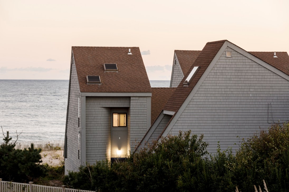 Modern house exterior with gray shingle siding, brown roof, skylights, and illuminated entrance near ocean