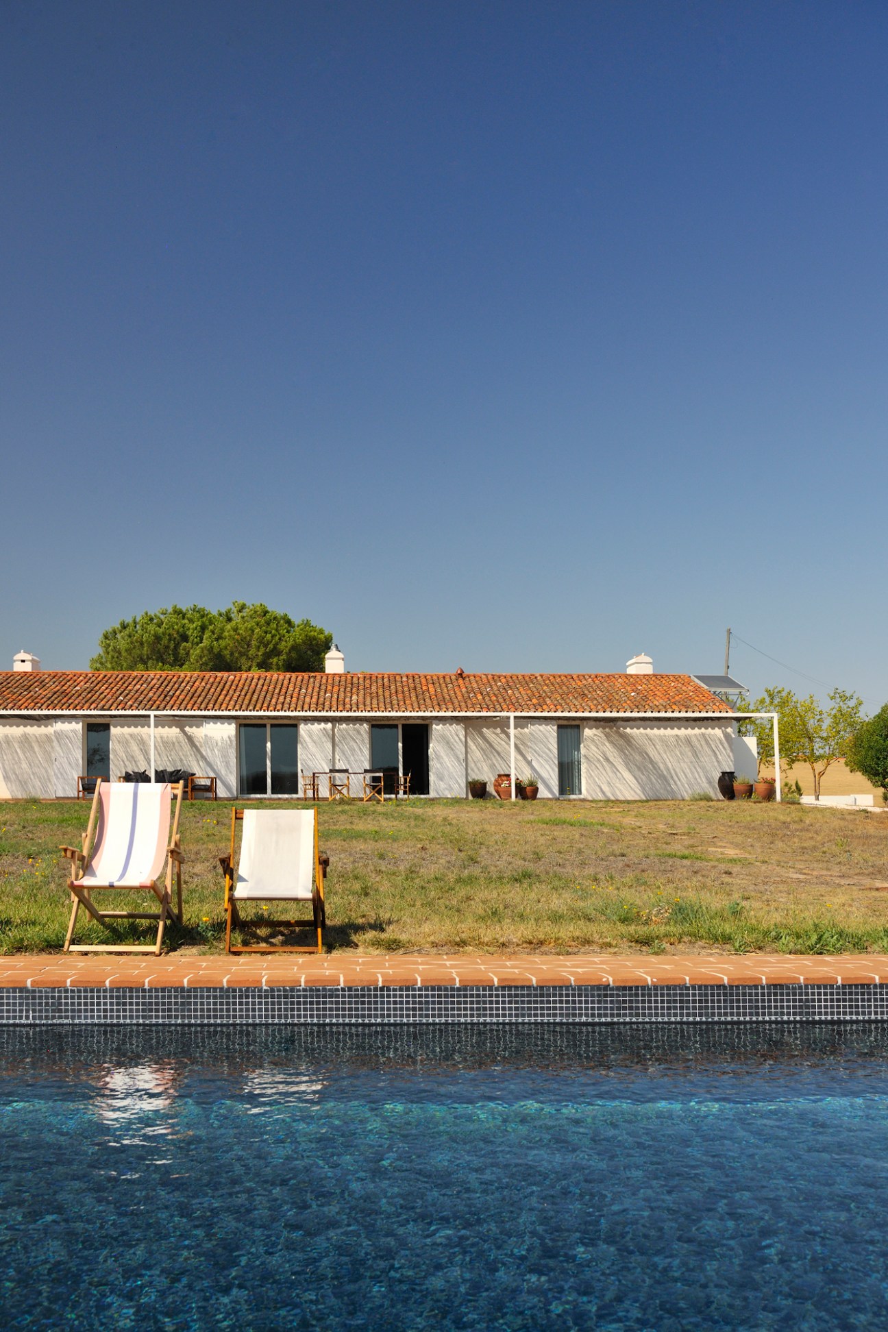 Modern single-story house with terracotta roof and white walls overlooking a swimming pool with two lounge chairs on deck