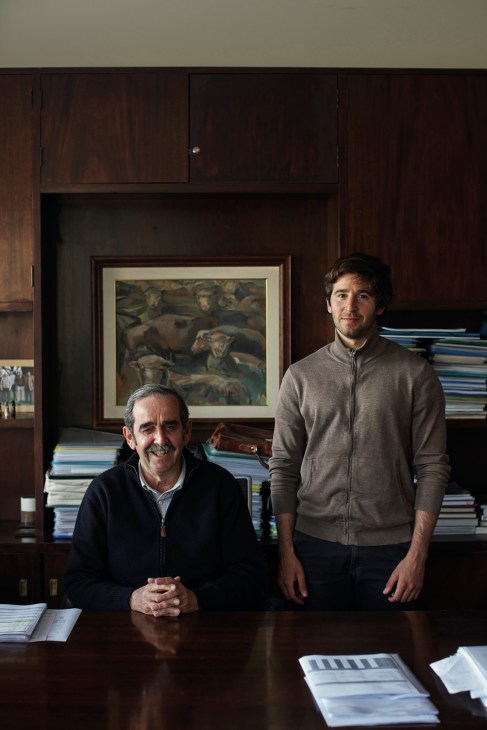 Two men pose in a wood-paneled office with stacked books and framed artwork behind them.