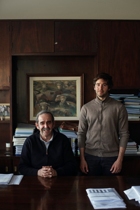 Two men pose in a wood-paneled office with stacked books and framed artwork behind them.