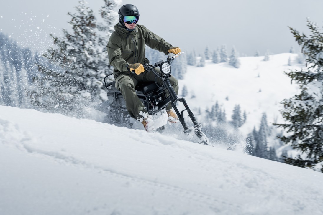 Person riding electric MoonBike through deep snow kicking up powder spray in mountain landscape