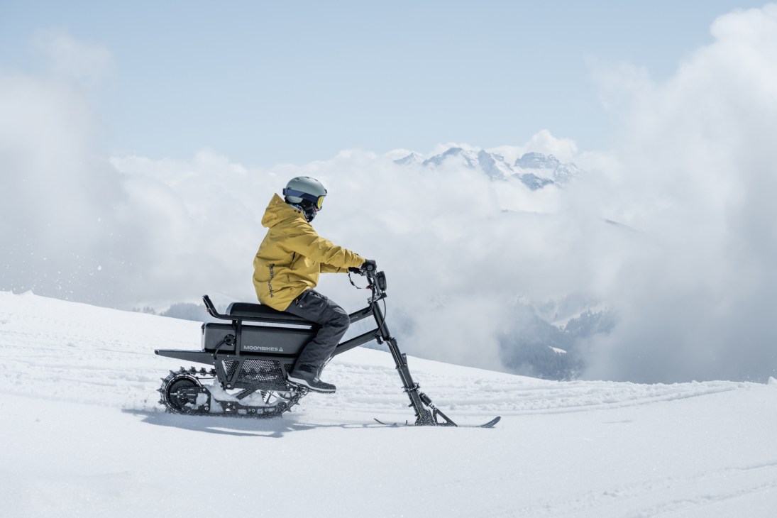 Person in yellow jacket riding a MoonBike electric snowbike across snowy mountain terrain