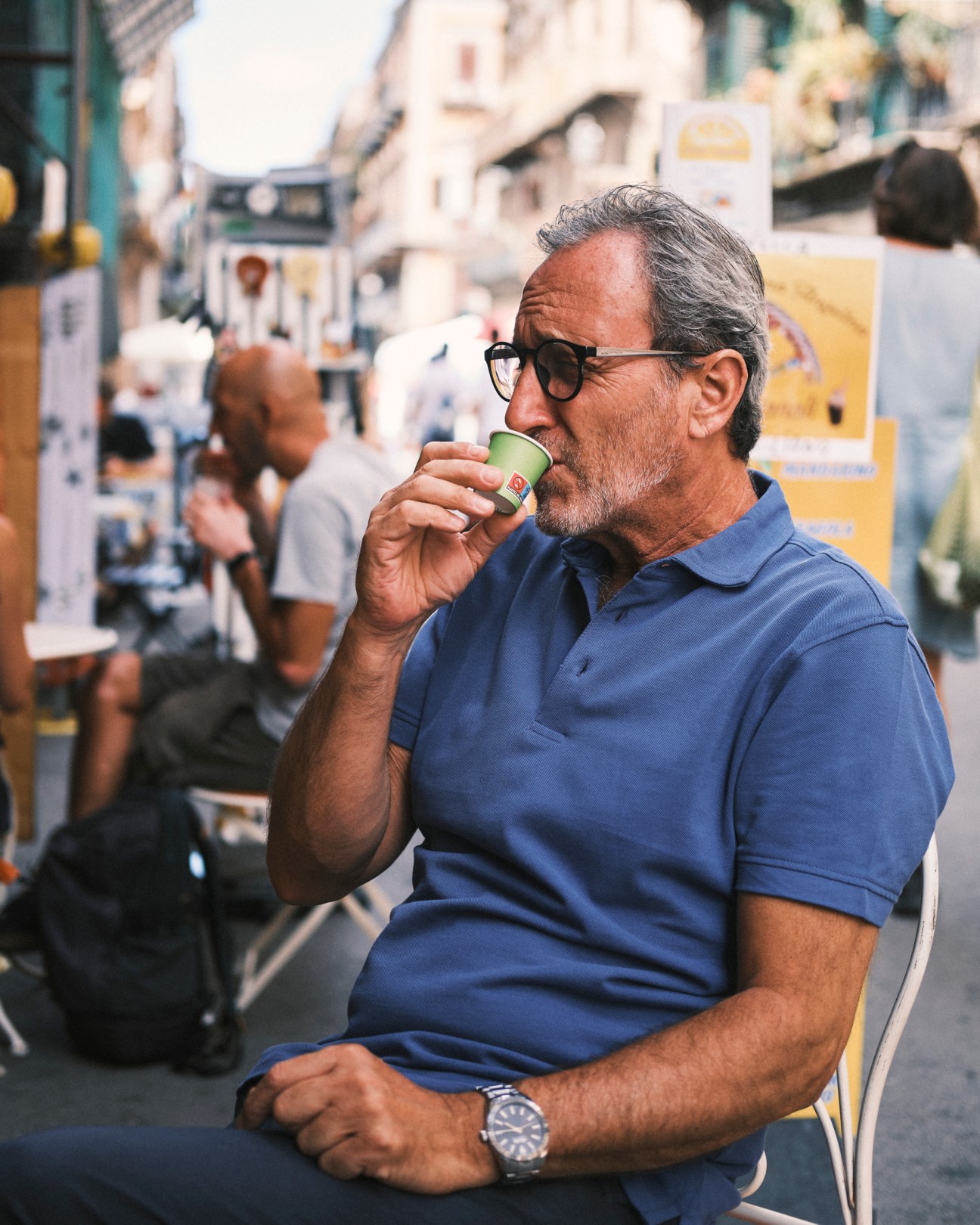 Man in sunglasses and blue polo shirt drinking espresso at outdoor café in Italian street setting