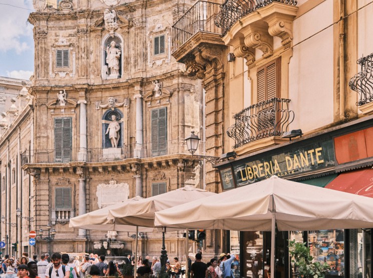Tourists and locals gather at outdoor cafe tables in historic Palermo square with baroque church architecture