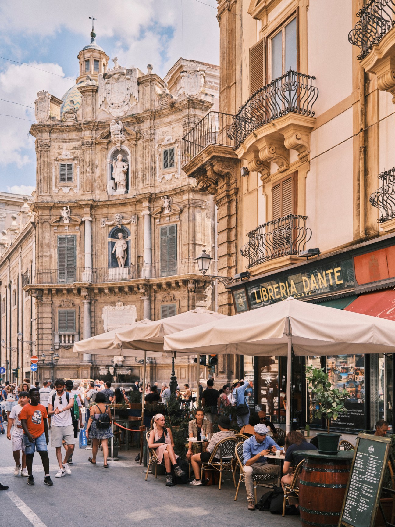 Tourists and locals gather at outdoor cafe tables in historic Palermo square with baroque church architecture