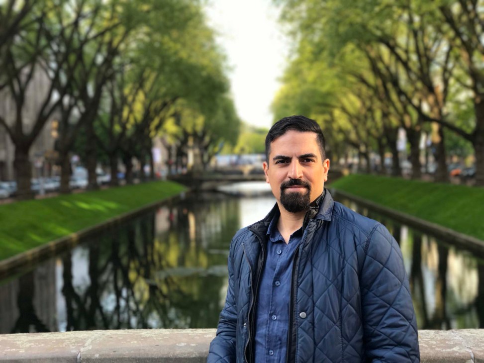 Portrait of a man with dark hair and beard wearing a dark blue quilted jacket in a tree-lined park with a canal.
