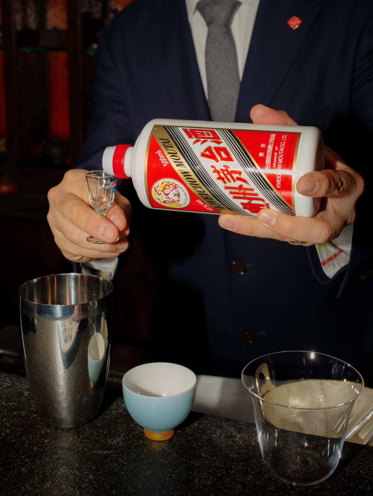 Person in suit pouring Kweichow Moutai baijiu from white bottle into small glass with ice bucket and cups nearby