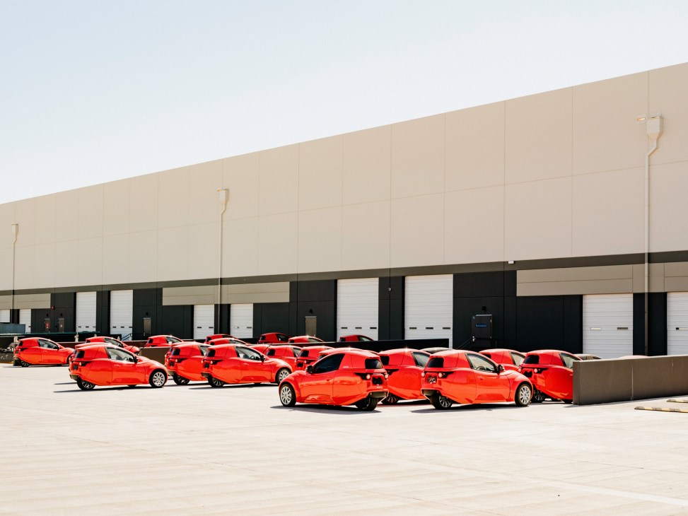 Row of orange Electrameccanica Solo electric vehicles parked outside industrial warehouse building