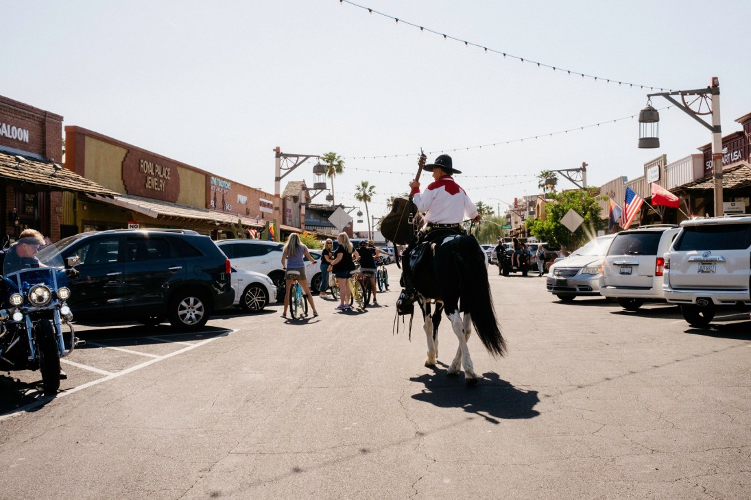 A person on horseback rides through a street lined with Old West-style buildings and parked cars