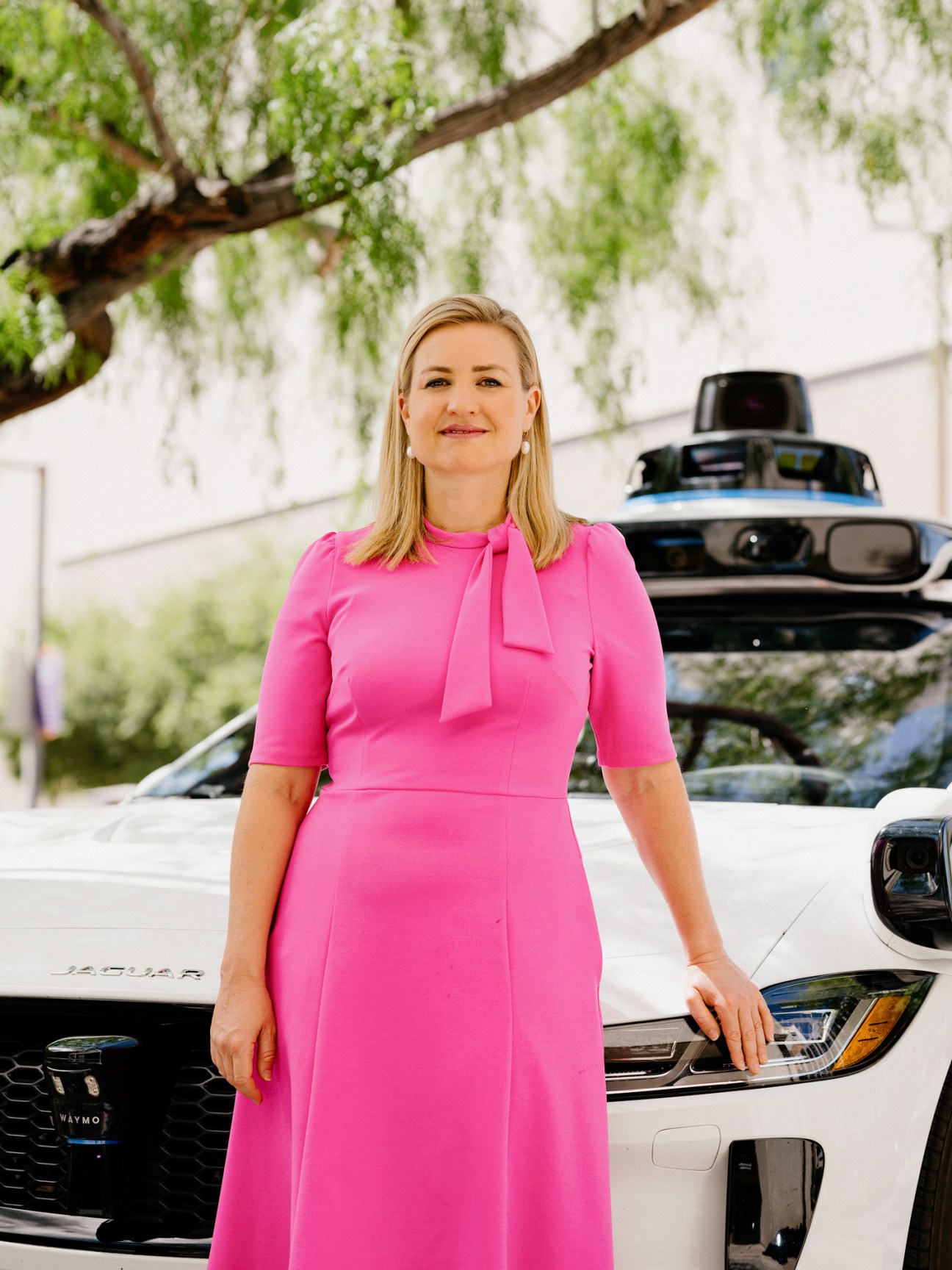 Phoenix mayor Kate Gallego in pink dress standing next to white Waymo autonomous vehicle with sensor equipment