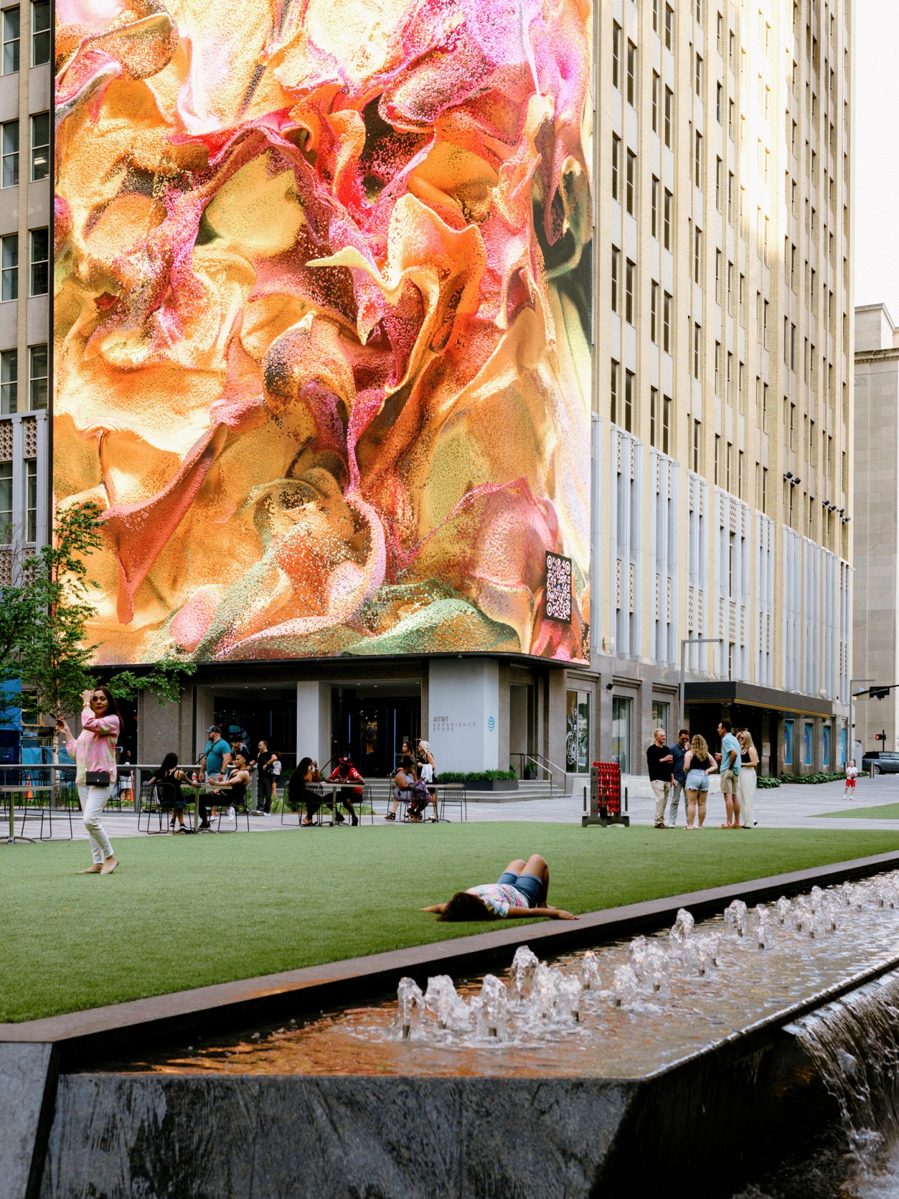 Large colorful mural on building facade above urban plaza with people, fountains and green space